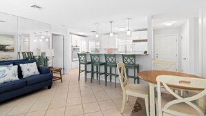 A modern living room with a blue sofa, a dining table with chairs, and a kitchen area with white cabinets and pendant lights. - Charming 2BR Condo Outrigger #4  by RVA (Longboat Key)