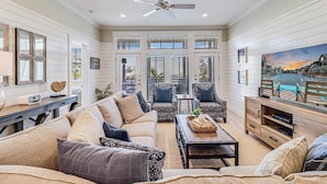 A spacious living room with a large sofa, two patterned armchairs, a coffee table, and a TV stand. There are multiple windows with shutters, a ceiling fan, and a painting of a coastal scene. - Custom, Designer Condo w/ Luxe Interiors+Enviable Amenities@Cinnamon Shore! (Port Aransas)