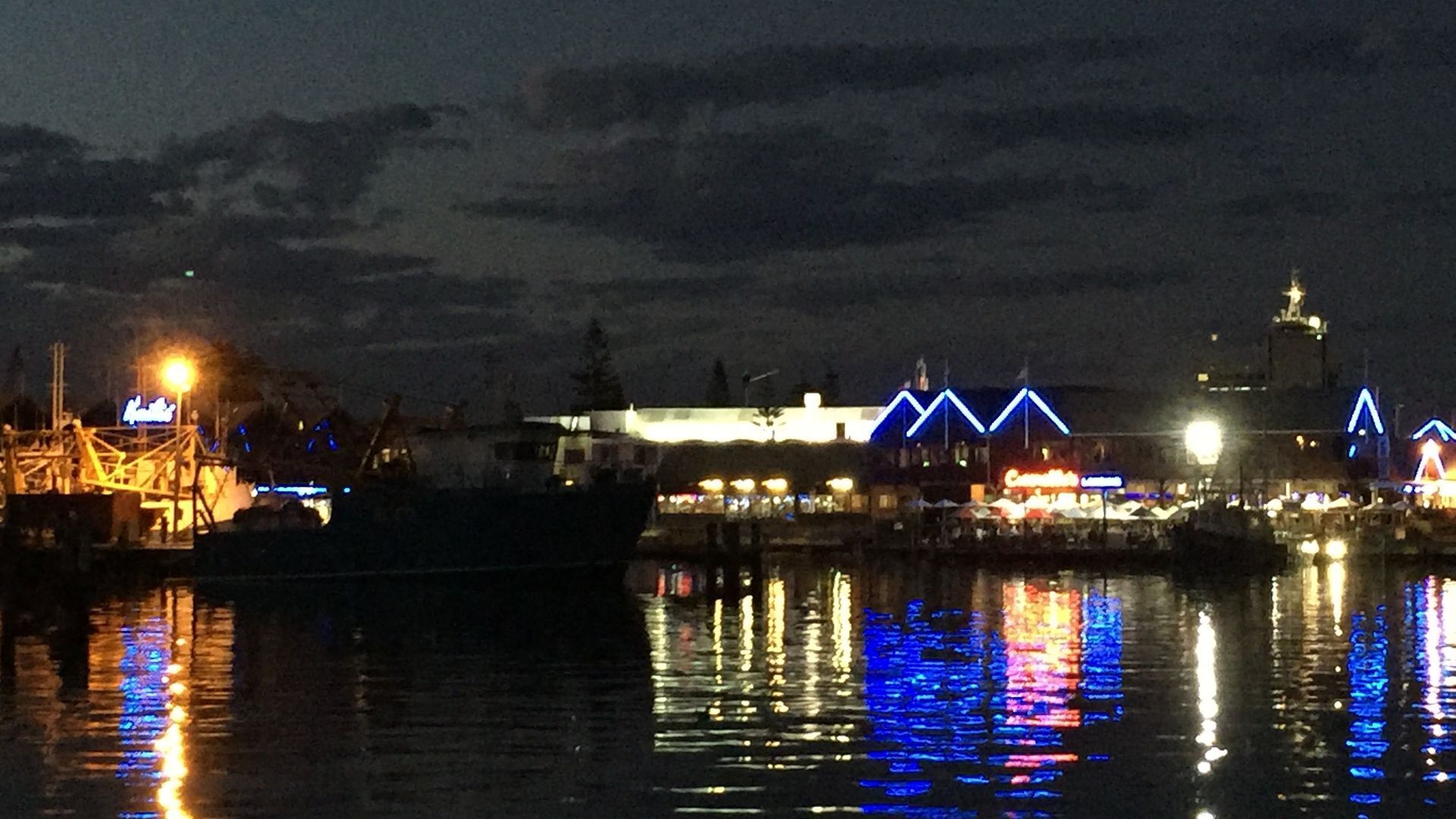 Un hôtel situé au bord de l’eau, avec des bâtiments illuminés de couleurs bleue et blanche, une zone abritant des quais et dont l’image se reflète sur l’eau.