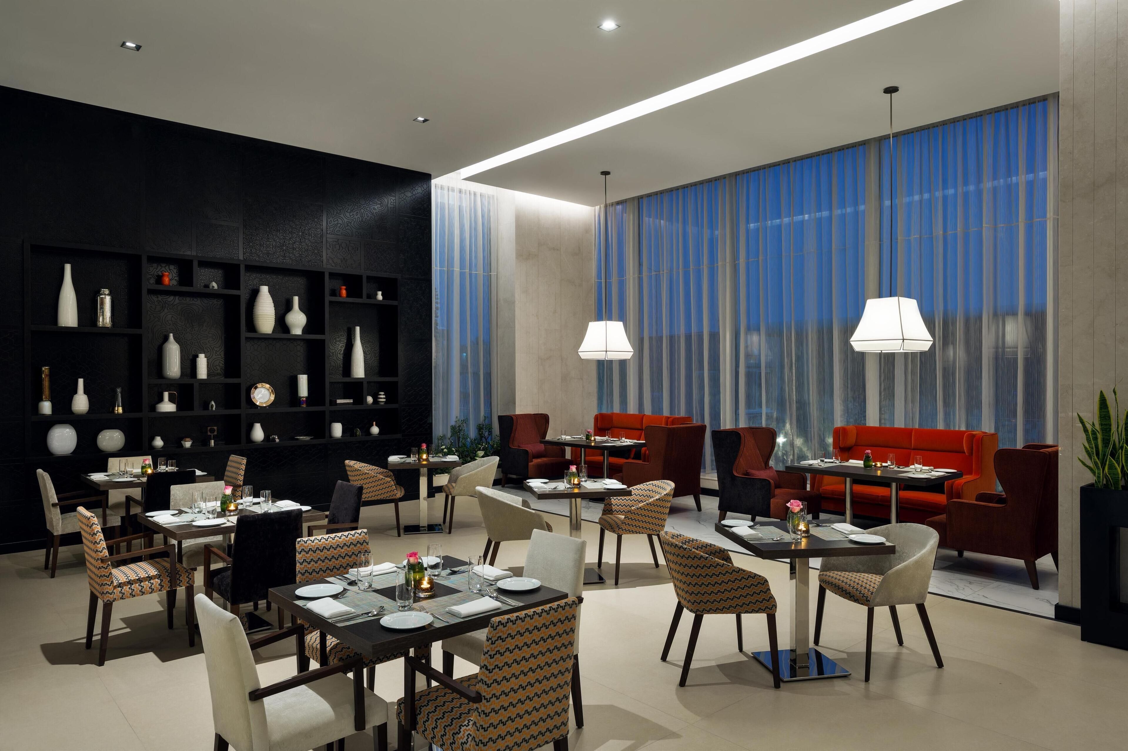 Un comedor moderno con mesas y sillas, una pared negra con estantes decorativos y ventanas amplias con cortinas azules.