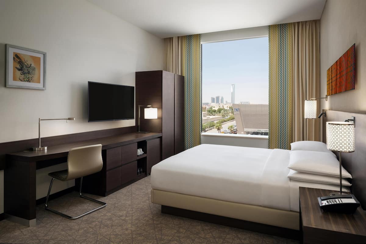 A hotel room with a bed, a desk, a chair, a television, and a window with a city view.