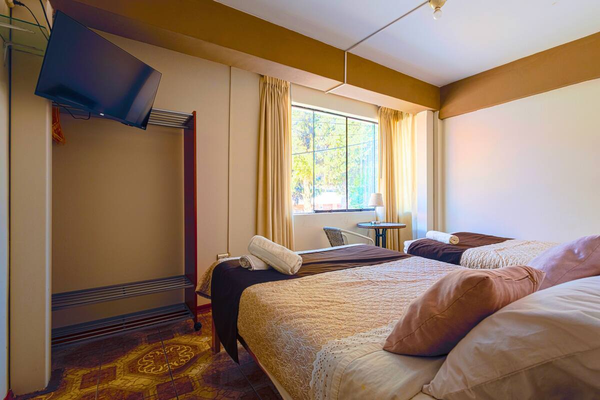 A hotel room with two beds, a television, a window with curtains, a small table, and a chair.