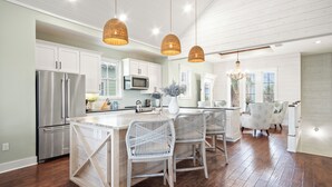 A spacious kitchen with white cabinetry, stainless steel appliances, wicker pendant lights, and a central island with chairs. - Pet-Friendly, Lakeside Home at Cinnamon Shore w/ Access to Pools & More (Port Aransas)