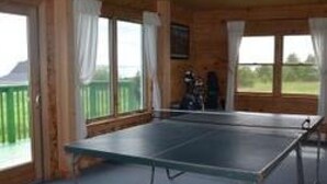 A room with a ping pong table, wooden walls, and large windows with curtains. - New Log Home with Gorgeous Ocean View in Historic Acadian Village (L'Ardoise)