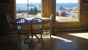 A cozy cabin with a wooden interior, a dining table with chairs, and large windows offering a view of the sea and a small building. - New Log Home with Gorgeous Ocean View in Historic Acadian Village (L'Ardoise)