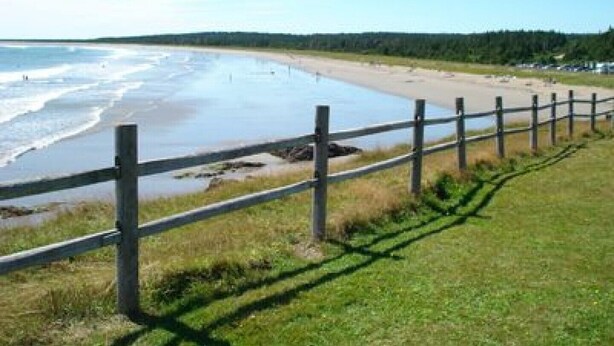 A beach with a wooden fence and grassy area in the foreground. - New Log Home with Gorgeous Ocean View in Historic Acadian Village (L'Ardoise)