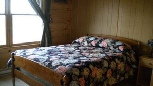 A wooden-framed bed with a floral-patterned bedspread in a room with wooden walls and a window with curtains. - New Log Home with Gorgeous Ocean View in Historic Acadian Village (L'Ardoise)