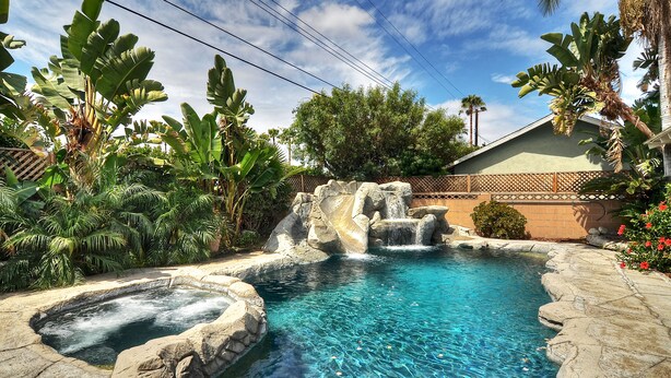 A swimming pool with a waterfall feature, surrounded by tropical plants and a brick wall. - **Water Slide!**Pool**Spa**Game Room!! (Anaheim)