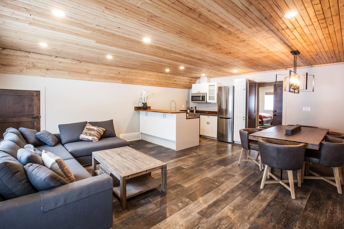 A modern interior with a wooden ceiling, a grey sofa, a wooden coffee table, a kitchen area with a fridge, microwave, and sink, and a dining table with chairs.