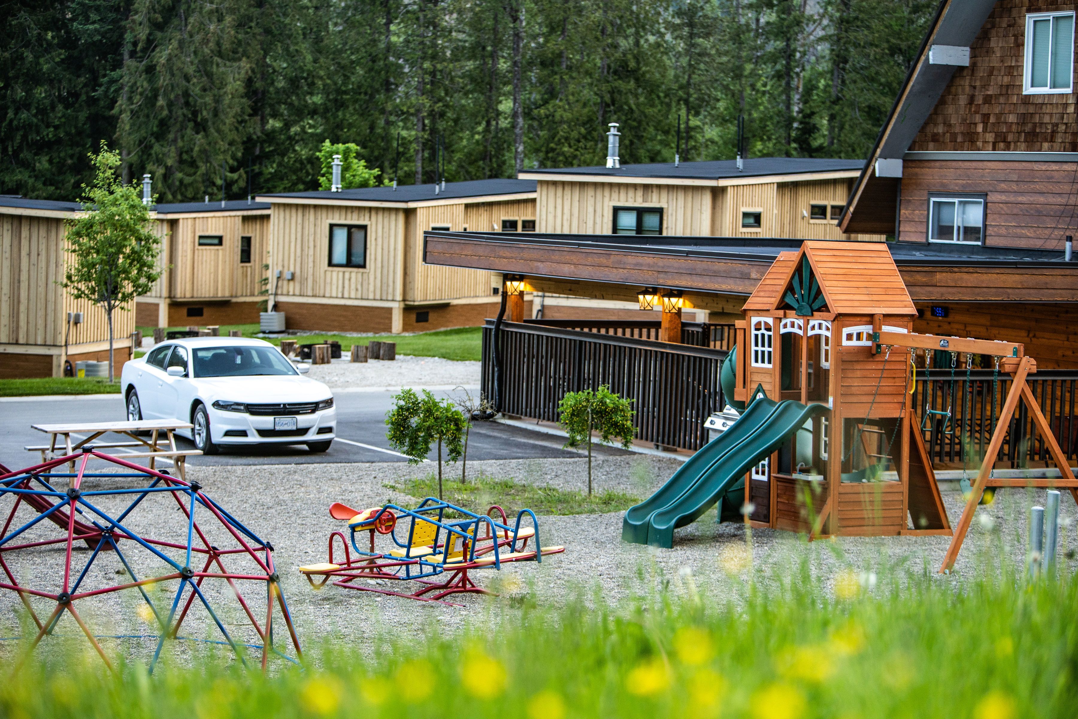Un terrain de jeux avec un toboggan et des balançoires, une voiture blanche garée à proximité, et un bâtiment à la façade en bois en arrière-plan.