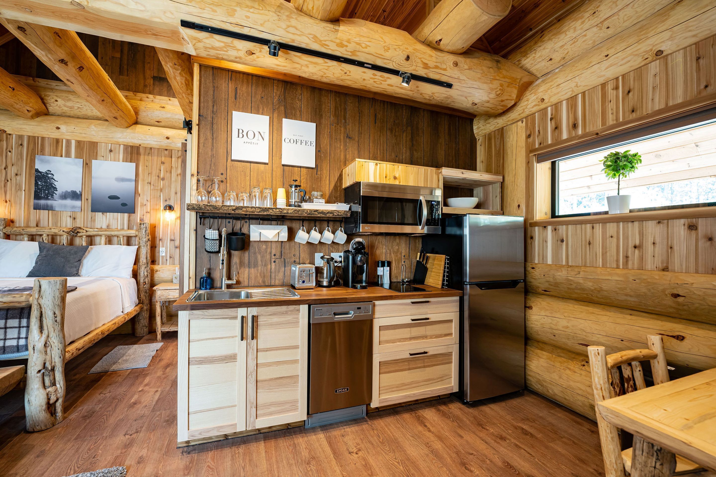 A rustic wooden cabin interior with a kitchen featuring wooden cabinets, a built-in microwave, and a refrigerator. There is a dining table and a bench with a view of the outdoors through a window.