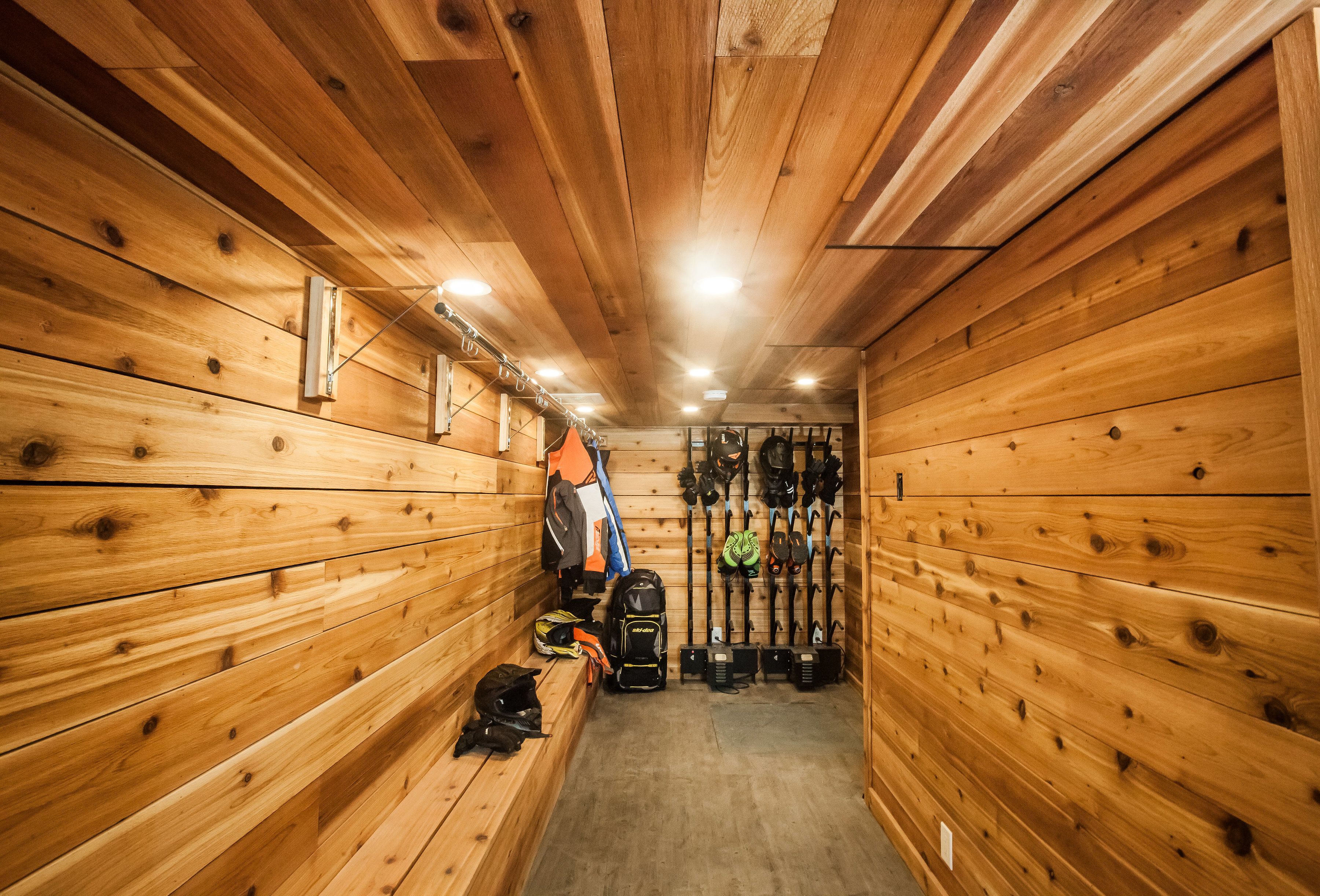 Una cabaña de madera con bancos a lo largo de las paredes, un perchero con esquíes y tablas de snowboard, y una lámpara en el techo.