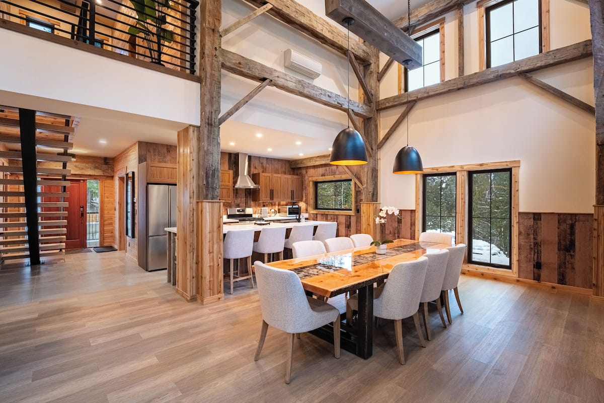 A spacious kitchen with wooden beams, a dining area with a long table and chairs, and large windows offering views of the outdoors.