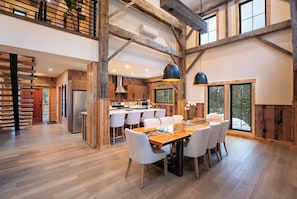 A spacious kitchen with wooden beams, a dining area with a long table and chairs, and large windows offering views of the outdoors. - Boulder Mountain Resort (Revelstoke)