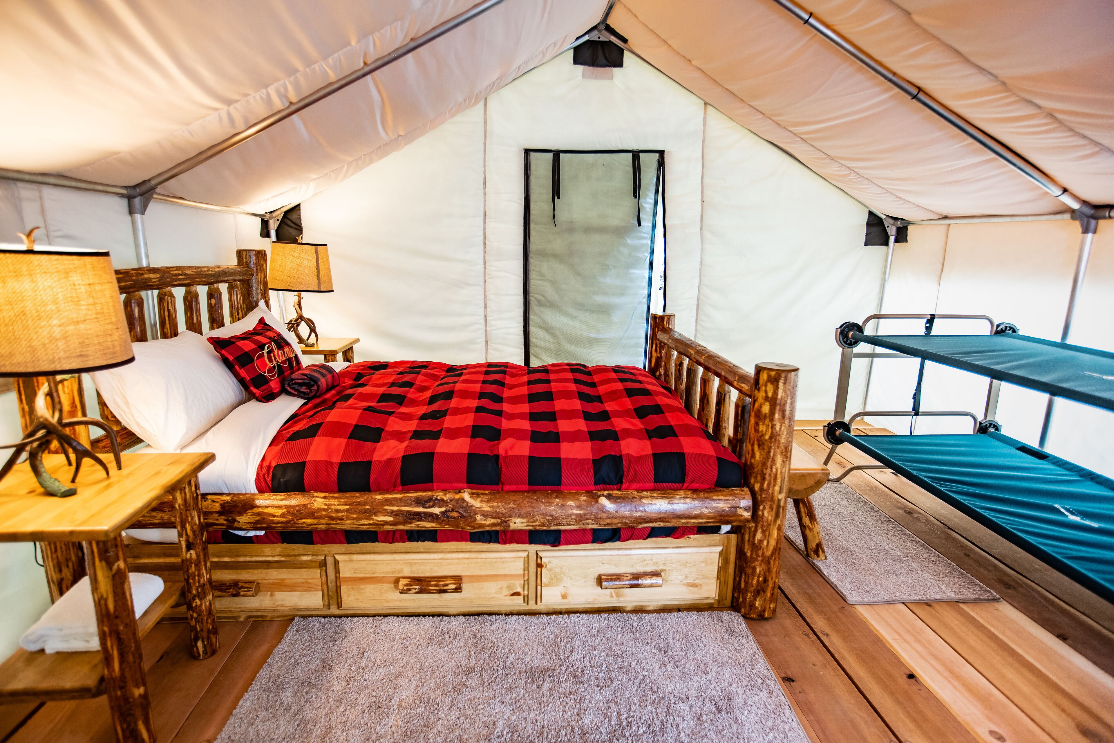 A tented room with a wooden bed, a small table with a lamp, and a bunk bed.