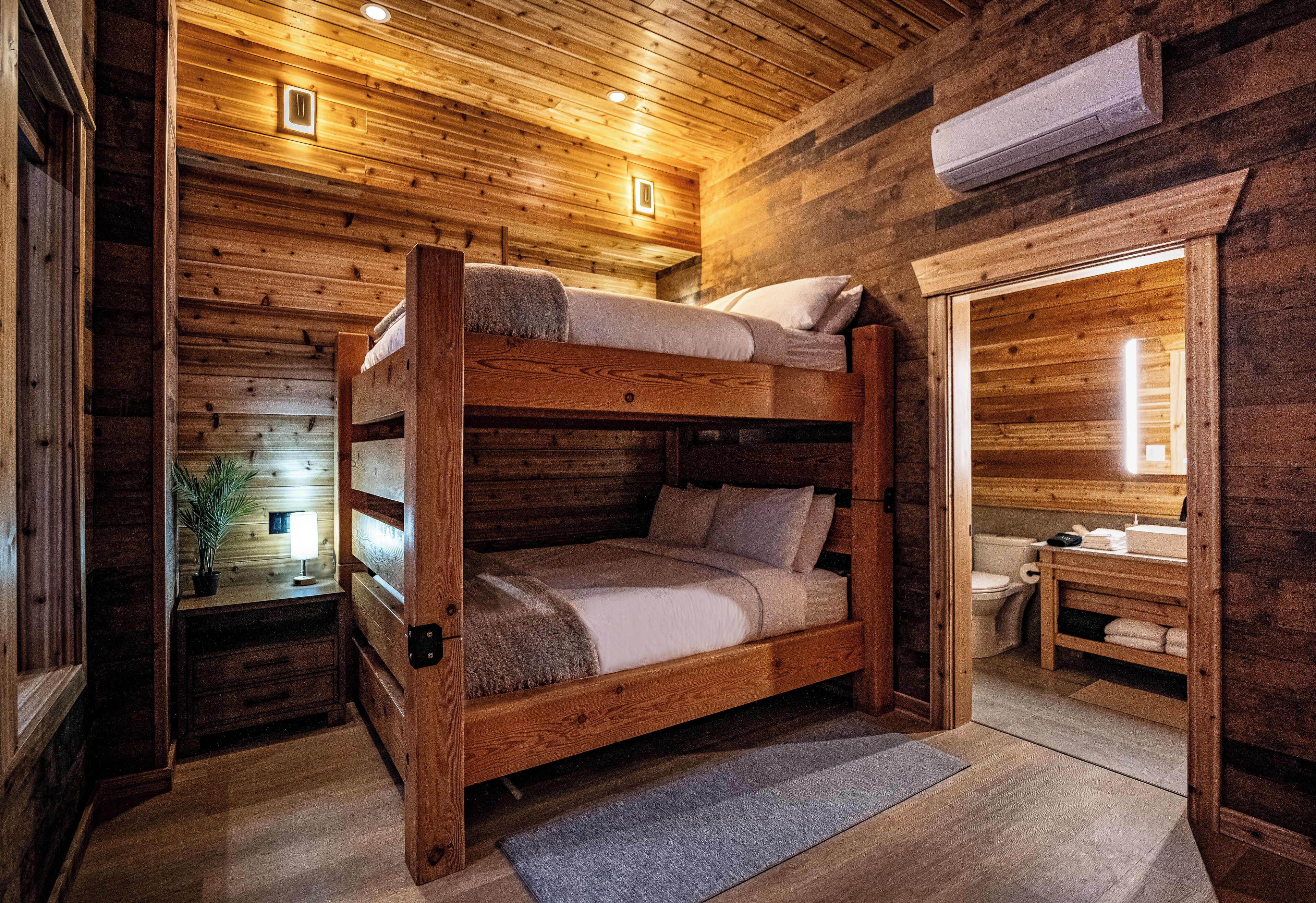 A wooden cabin with a bunk bed, a small bedside table with a lamp, and a bathroom with a toilet and sink.
