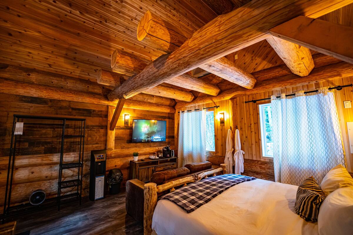 A log cabin room with a bed, a TV, a desk, and a window with curtains.