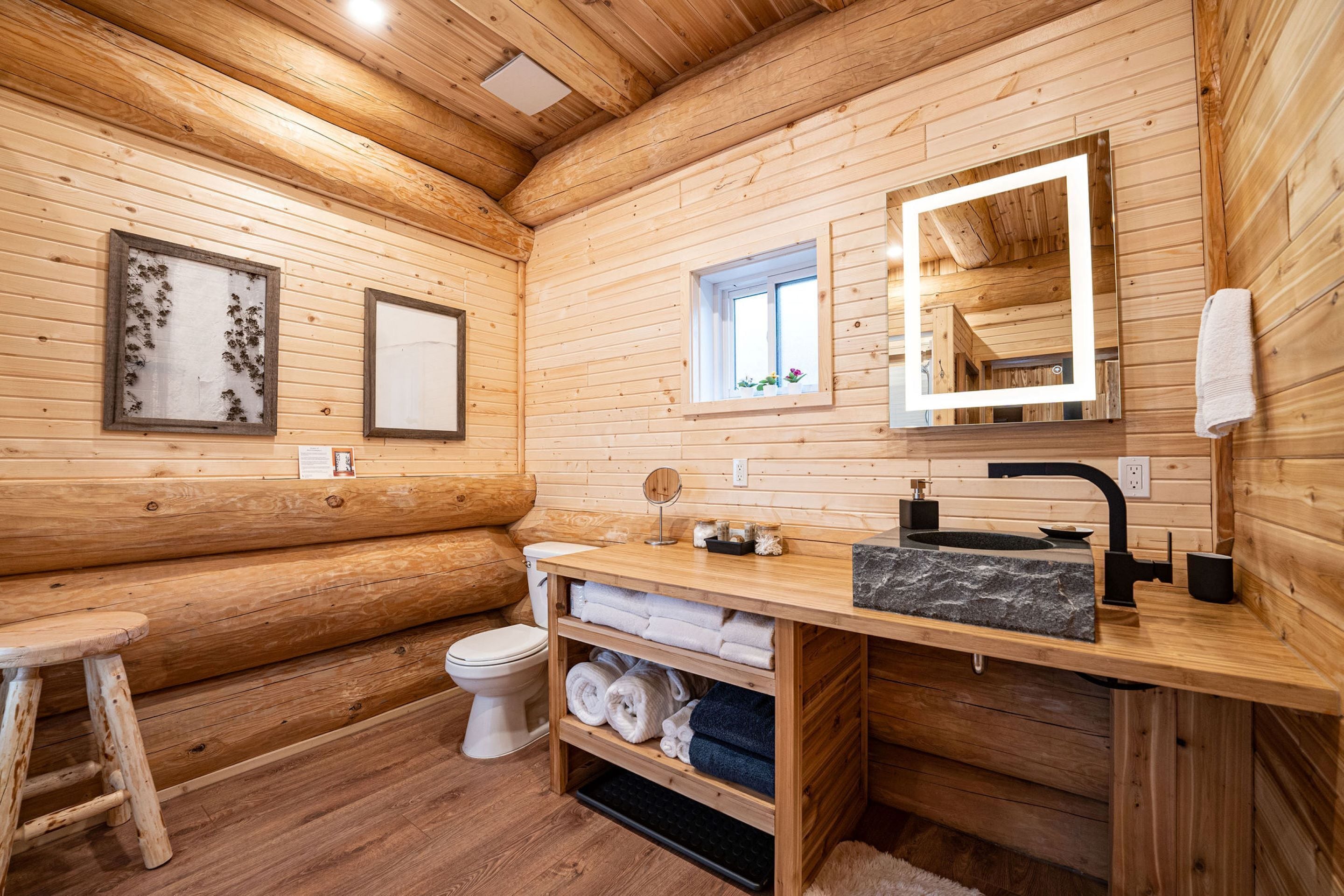 A wooden cabin bathroom with a wooden bench, toilet, sink with a marble countertop, and a mirror with a light.