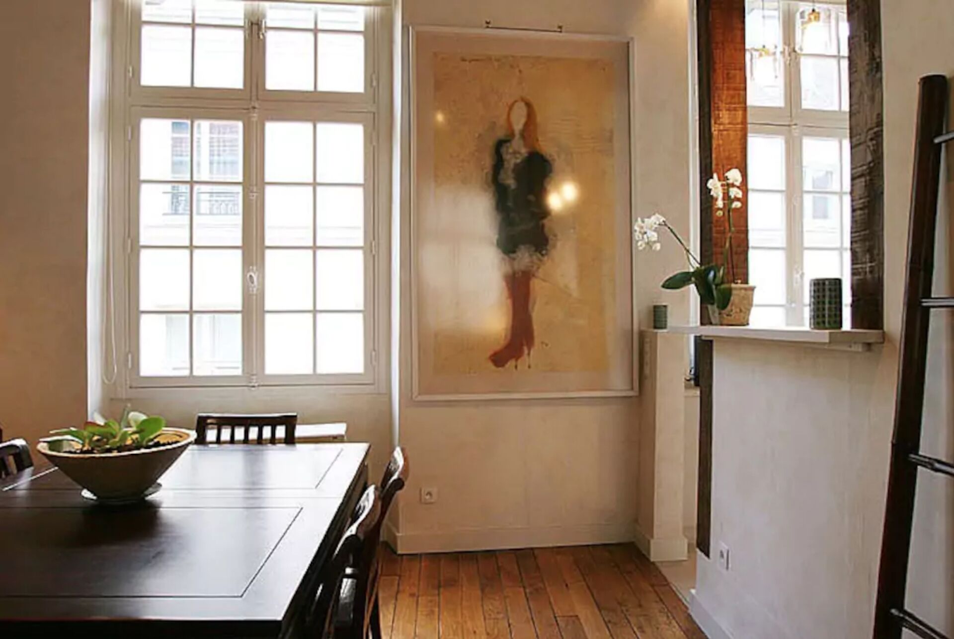 Un comedor con una mesa de madera, sillas, una ventana con persianas, una obra de arte en la pared y una planta en maceta.