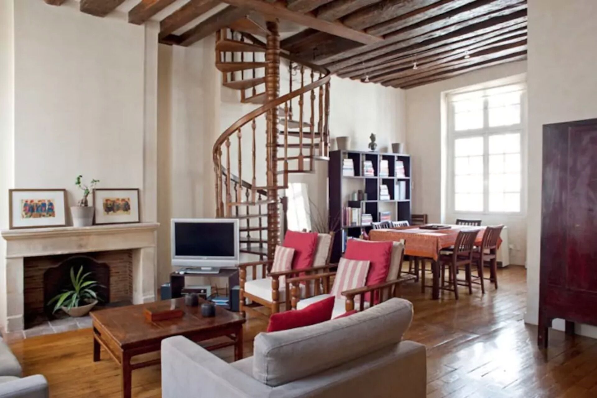 A spacious living room with a wooden staircase, a fireplace, a dining area, and a bookshelf.