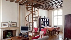 A spacious living room with a wooden staircase, a fireplace, a dining area, and a bookshelf. - Charming cozy triplex in trendy Marais (Paris)