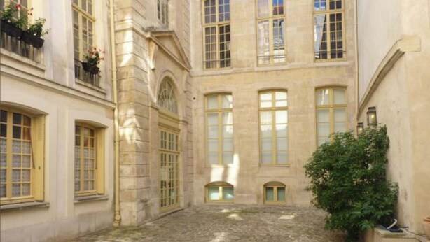 A courtyard with cobblestone flooring, large windows with yellow frames, and a potted plant. - Charming cozy triplex in trendy Marais (Paris)
