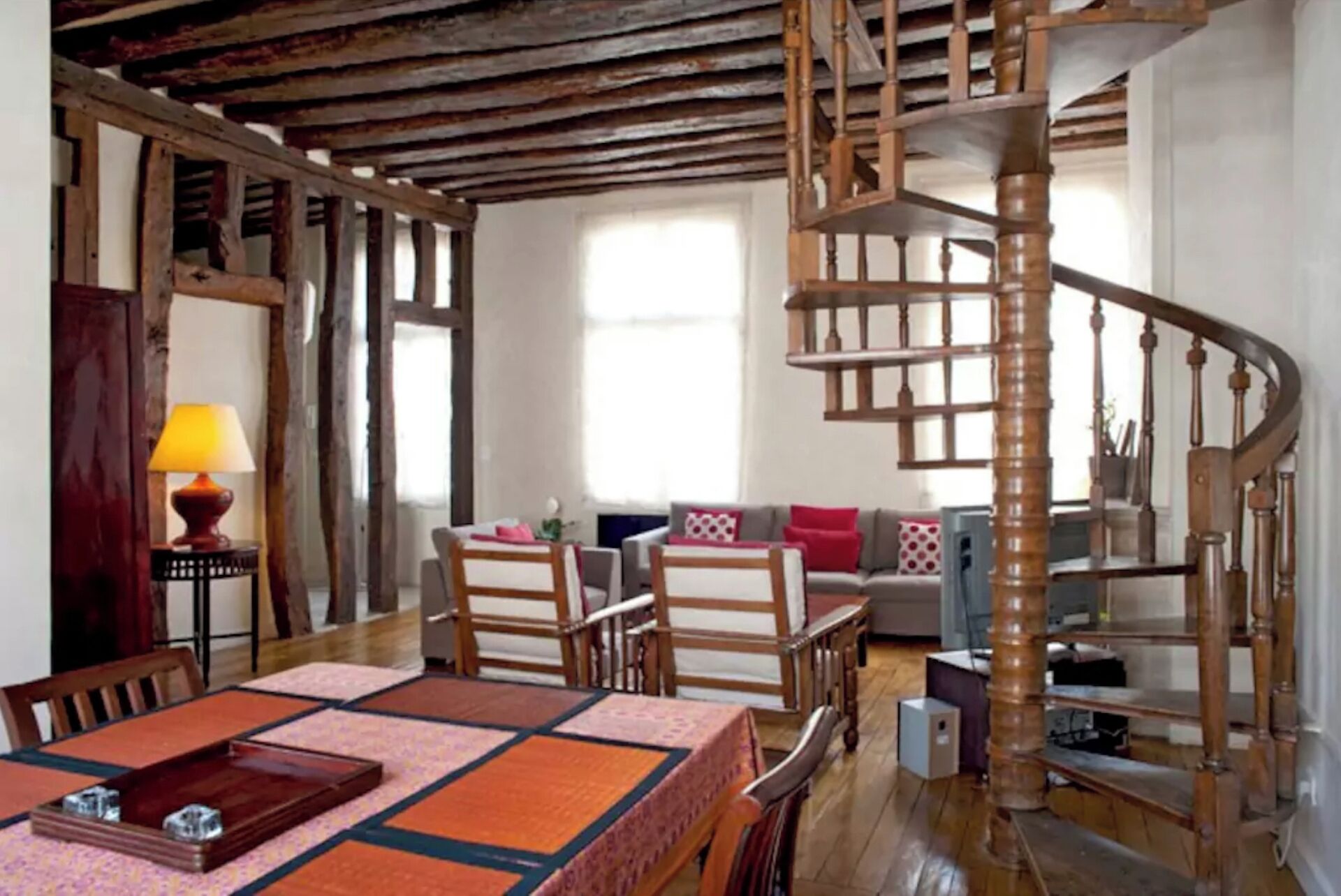 A rustic interior with exposed wooden beams, a wooden spiral staircase, a dining area with a table and chairs, and a sofa with cushions.