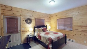 A bedroom with wooden walls, a bed with a quilt, two bedside lamps, a nightstand, a door, and a window with blinds. - 31 MI to YNP | Fire Pit | Sleeps 10 (Island Park)