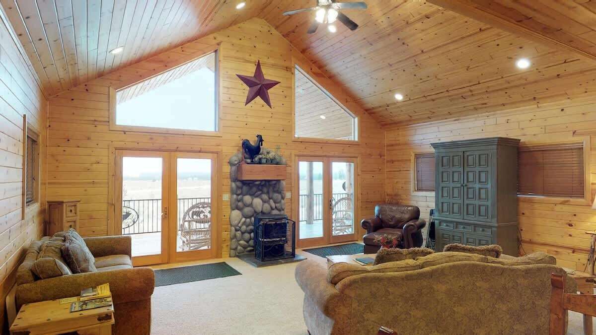 A spacious log cabin interior with a stone fireplace, two sofas, a ceiling fan, and large windows offering views of the outdoors.