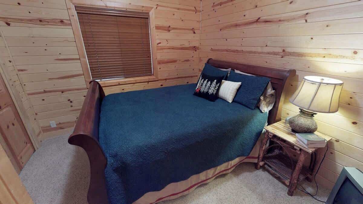 A wooden cabin bedroom with a bed, a nightstand, a lamp, and a window with blinds.
