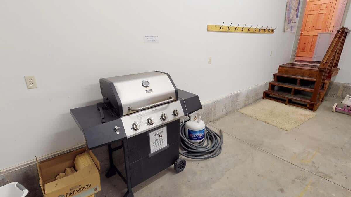 A portable grill with a gas tank and a box of firewood in a room with a concrete floor and a wooden staircase.