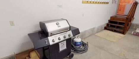 A portable grill with a gas tank and a box of firewood in a room with a concrete floor and a wooden staircase.