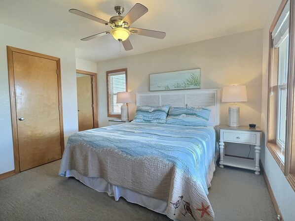 A bedroom with a bed, a nightstand, a lamp, a ceiling fan, a door, a window with blinds, and a wall-mounted artwork. - Shell Castle Cottage- 4 Bdrm- Steps from beach- Community Pool & Spa (Buxton)
