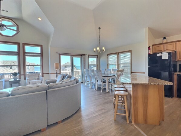 Amplia sala de estar con un sofá grande, una mesa de comedor y sillas, una cocina con refrigerador y armarios de madera, y ventanas amplias que ofrecen vistas al mar.