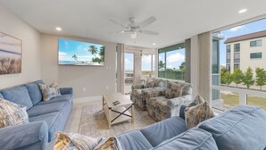 A spacious living room with a large sofa, a smaller sofa, and a coffee table. There is a ceiling fan, a large window with a view of palm trees, and a balcony with outdoor furniture.
