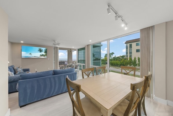 Un salon moderne avec un canapé bleu, une table à manger en bois et un balcon offrant une vue sur l’océan et les bâtiments.