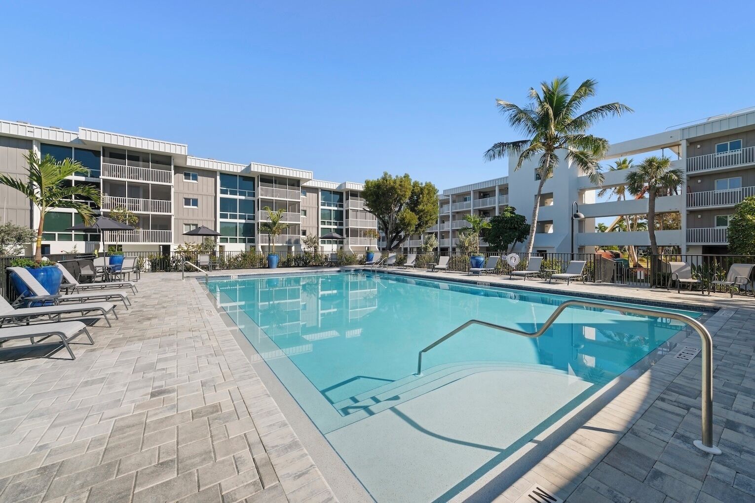 A swimming pool with lounge chairs and a brick patio, surrounded by multi-story buildings with balconies and palm trees.