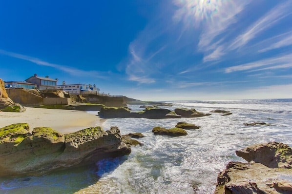 A coastal scene with a building on the left, rocky shoreline, and waves crashing onto the beach.