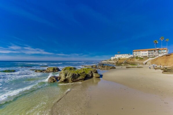Una playa con cielos azules despejados, costa rocosa y un edificio con techo rojo.