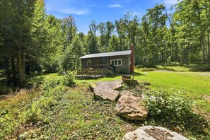 Uma cabana com varanda, rodeada por uma floresta e um espaço gramado.