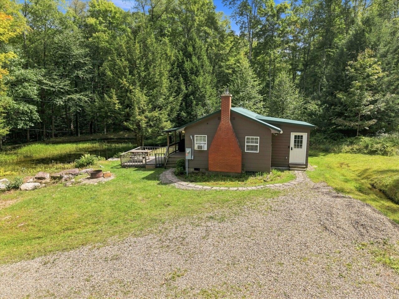 A small cabin with a brick chimney, surrounded by a gravel driveway and a grassy area.