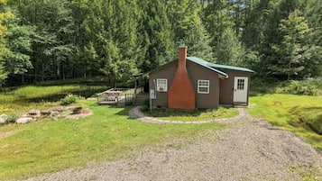 A small cabin with a brick chimney, surrounded by a gravel driveway and a grassy area.