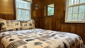 Dormitorio de cabaña de madera con una cama grande, ropa de cama escocesa y varias ventanas con vistas al bosque.