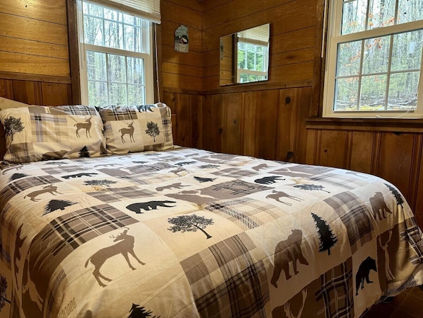 Ein Schlafzimmer einer Holzhütte mit einem großen Bett, Karo-Bettwäsche und mehreren Fenstern mit Blick auf einen Wald.