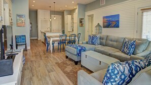 Un salon spacieux avec un canapé gris, des coussins à motifs bleus, un coin repas avec une table blanche et des chaises bleues, et une cuisine en arrière-plan.