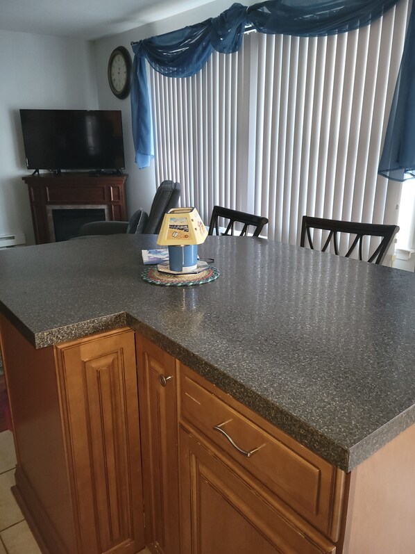 A kitchen with a granite countertop, wooden cabinets, a small island, a clock on the wall, and a TV above the fireplace.