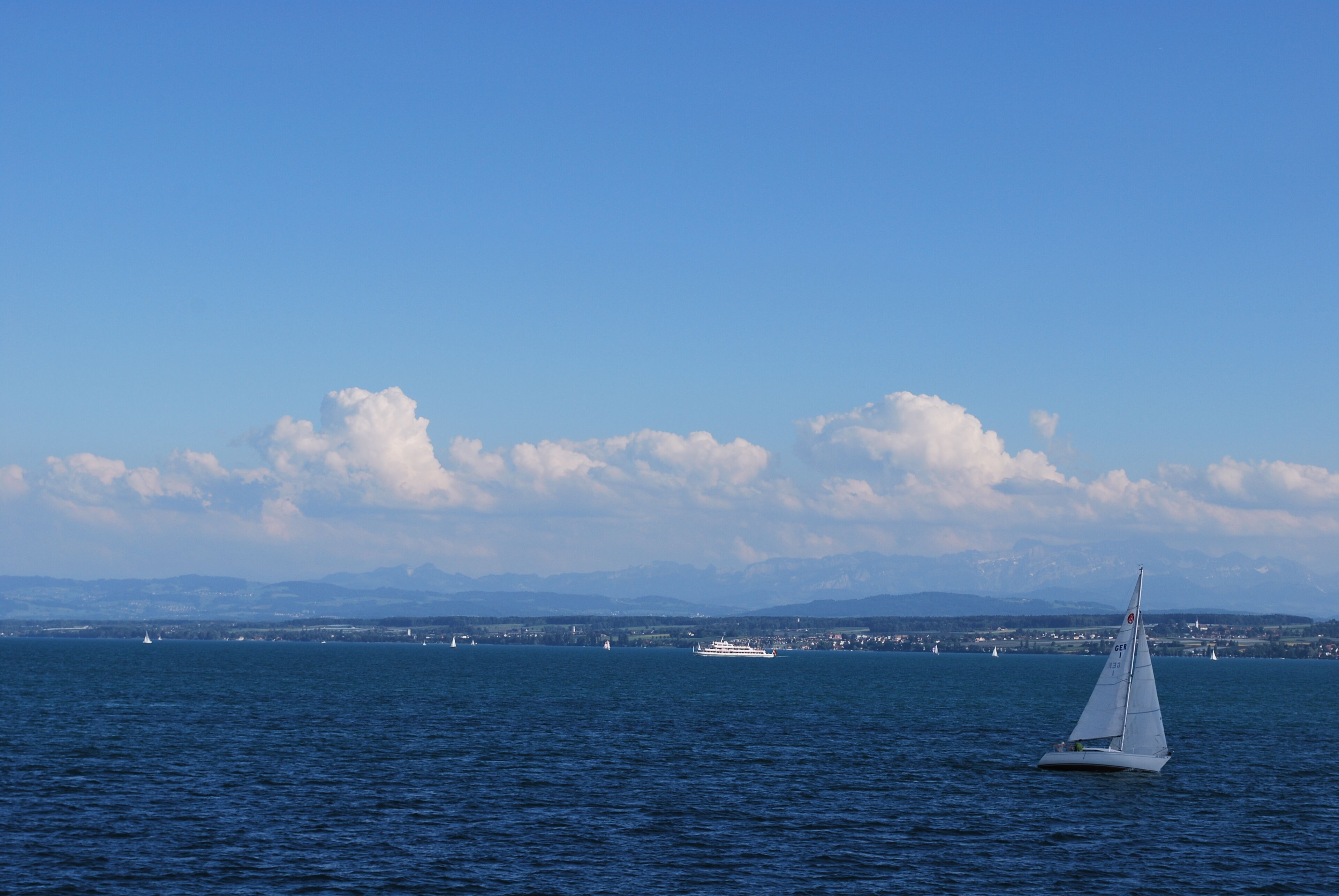 Un velero en aguas tranquilas, con un paisaje urbano distante y montañas al fondo.