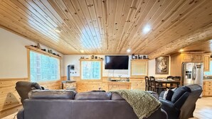 Un salon chaleureux avec un grand canapé gris, un téléviseur à écran plat, un plafond en bois et un coin repas comprenant une table et des chaises.