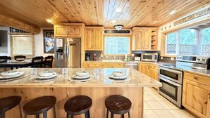Une cuisine spacieuse avec des meubles en bois, un grand îlot central doté de tabourets, des appareils en acier inoxydable et une vue sur l’extérieur.