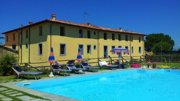 Ein Poolbereich mit Liegestühlen und Sonnenschirmen, ein gelbes Gebäude mit einem Ziegeldach und einem strahlend blauen Himmel.